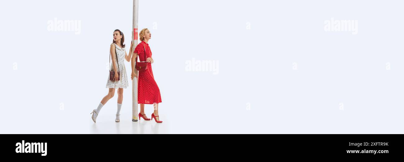 Due donne vestite con eleganti abiti a tema retrò che trasportano un tubo di cartone gigante isolato su sfondo bianco. Designer di interni. Foto Stock