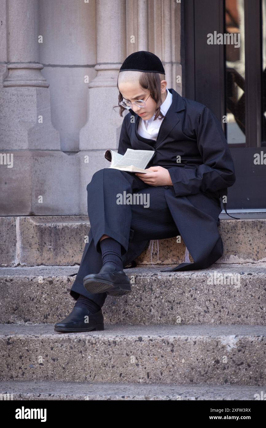 Durante la pausa, un giovane ebreo ortodosso studioso siede sui gradini della scuola e studia il Talmud. A Brooklyn, New York, marzo 2024. Foto Stock