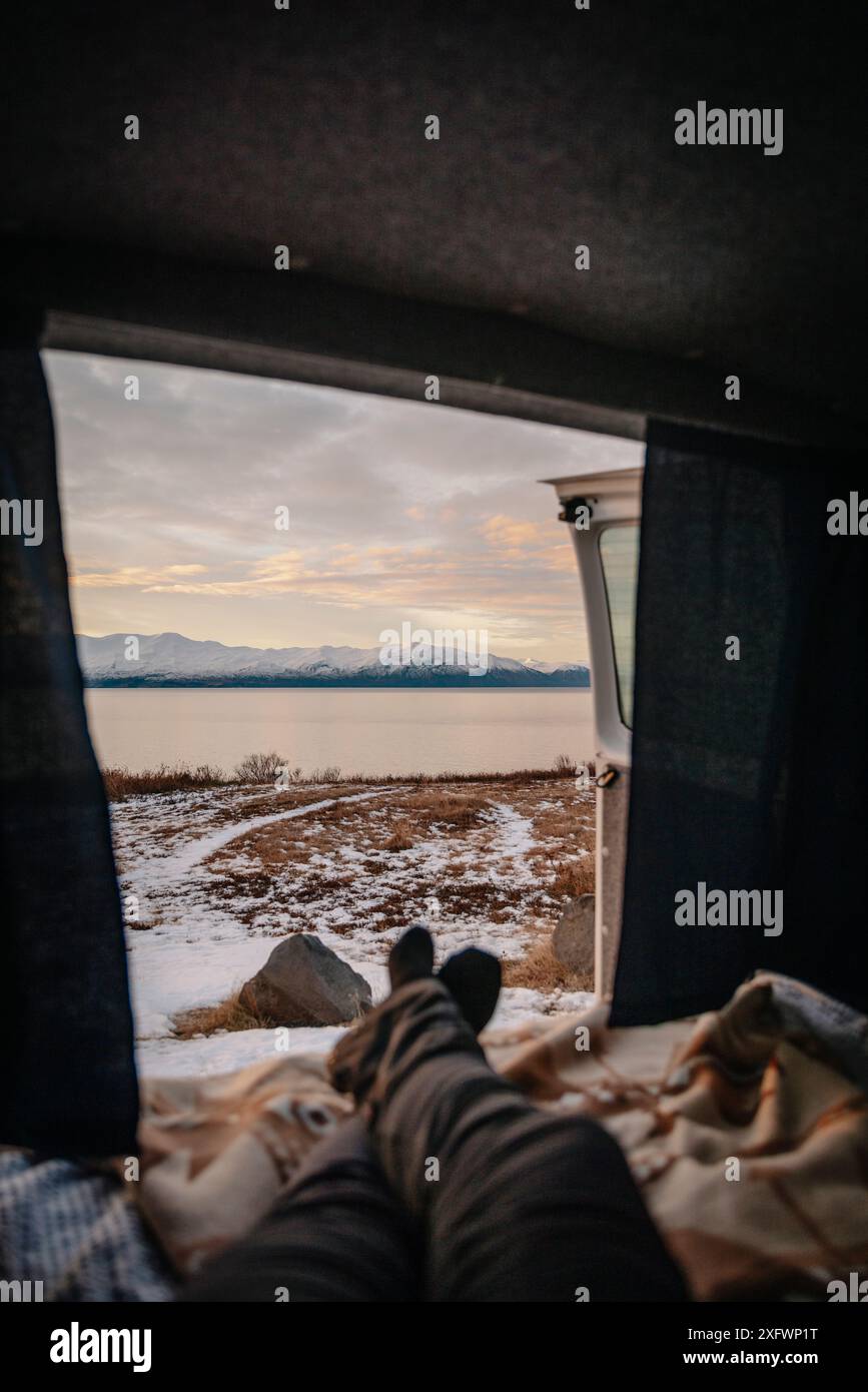 Parte bassa della persona che si rilassa all'interno del camper vicino al mare e alle montagne al tramonto Foto Stock