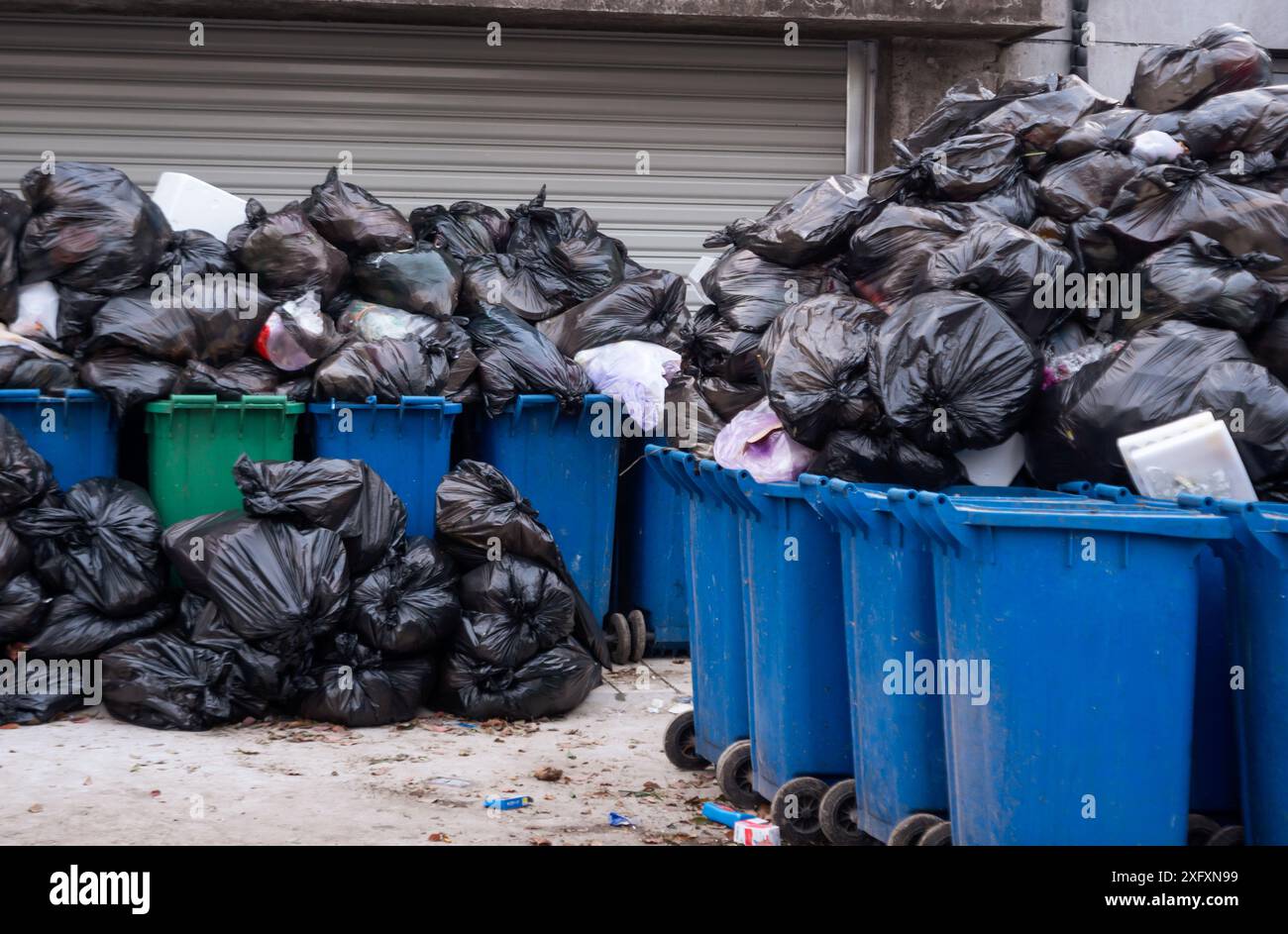 Una pila di sacchetti neri traboccanti che si riversano sulla sommità di diversi contenitori di riciclaggio blu e verdi in una strada cittadina. Traboccamento attivo Foto Stock