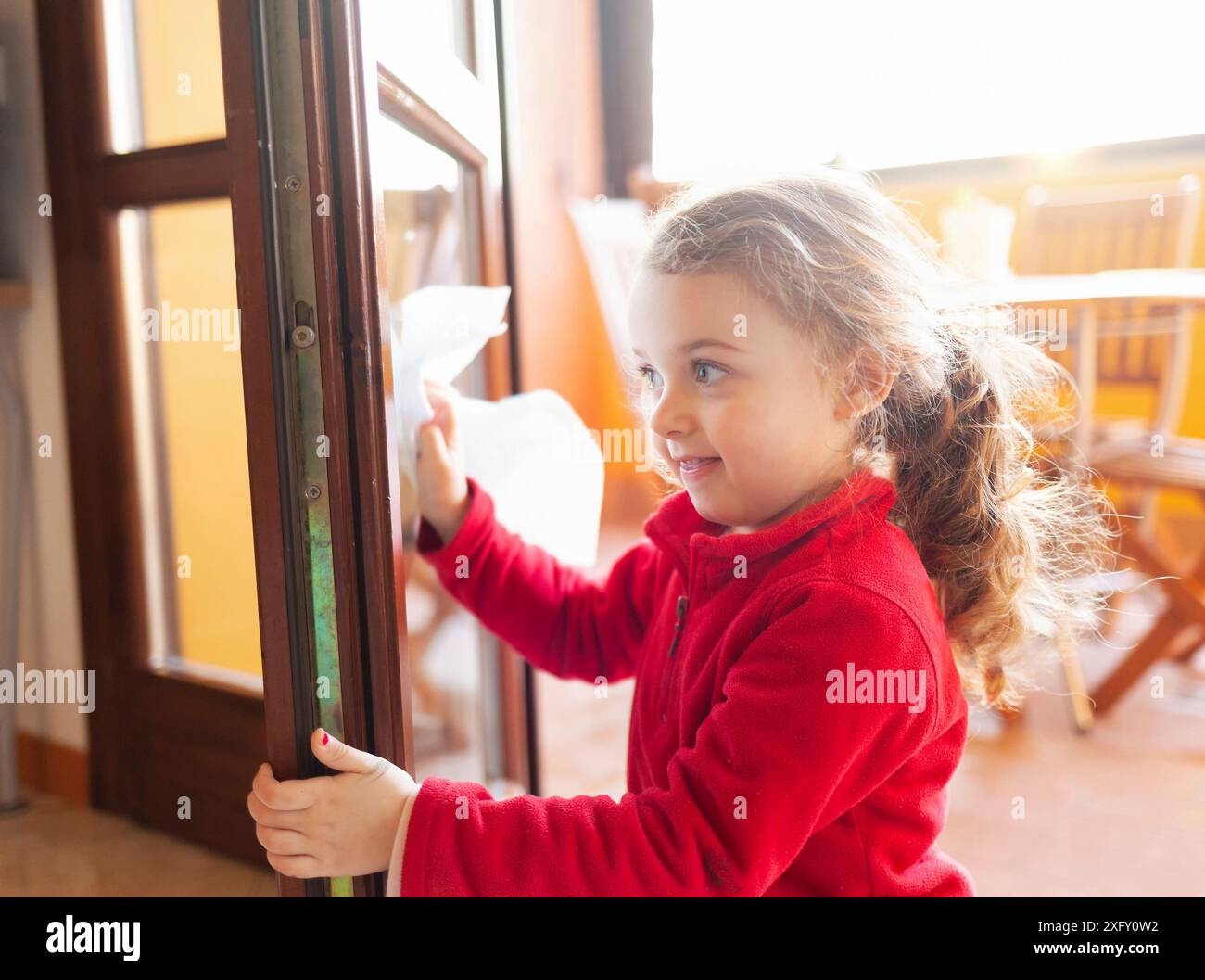 La ragazza di tre anni aiuta con la pulizia della famiglia. Pulisce le finestre con un foglio di carta. Idea di passare il tempo durante il periodo di quarantena del coronavirus covid-19. Foto Stock