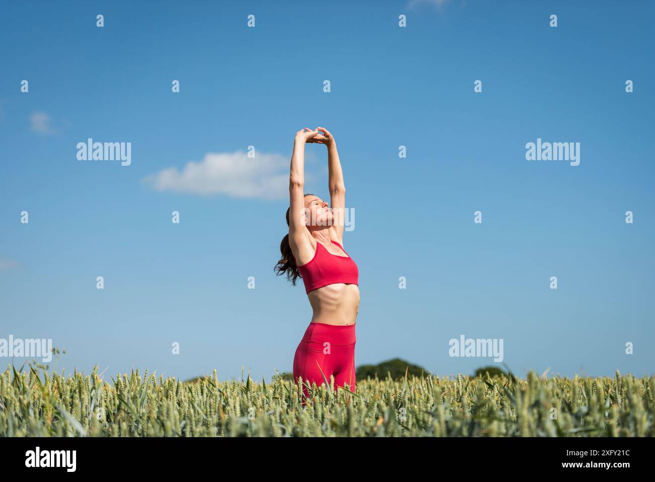 Donna sportiva con le braccia sollevate che fa esercizi di stretching nel campo del grano, indossa abbigliamento sportivo rosso con il cielo blu. Foto Stock