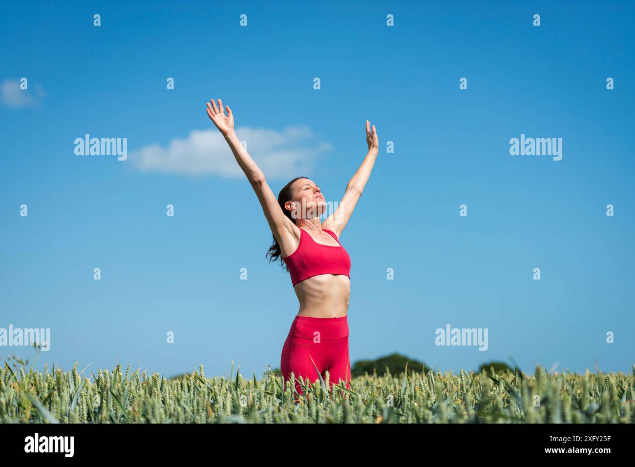 Donna sportiva con le braccia alzate per festeggiare e godersi il sole nel campo del grano, indossando abbigliamento sportivo rosso con il cielo blu. Foto Stock