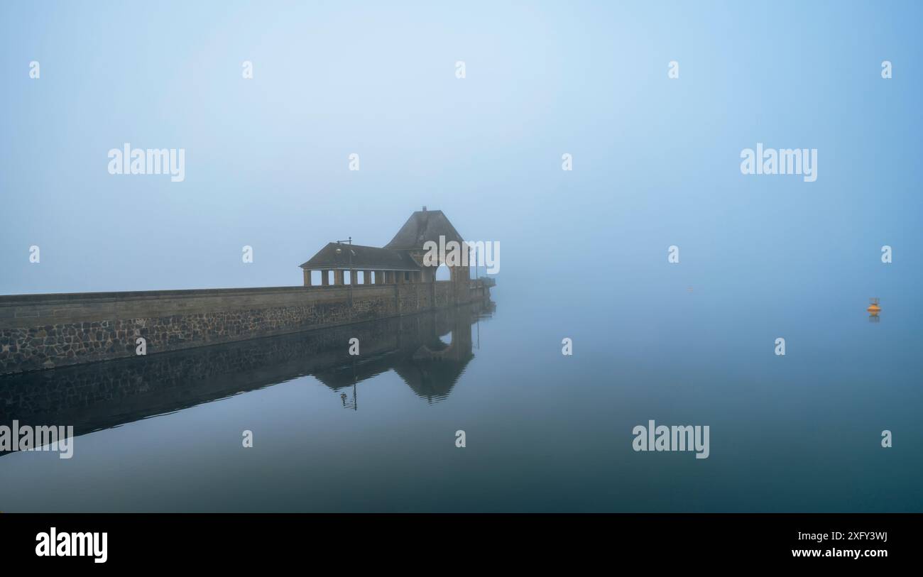 Parete della diga sul lago Edersee nella nebbia mattutina, riflesso nelle acque calme, una boa gialla sul bordo della foto. Distretto di Waldeck-Frankenberg, Assia, Germania. Foto Stock