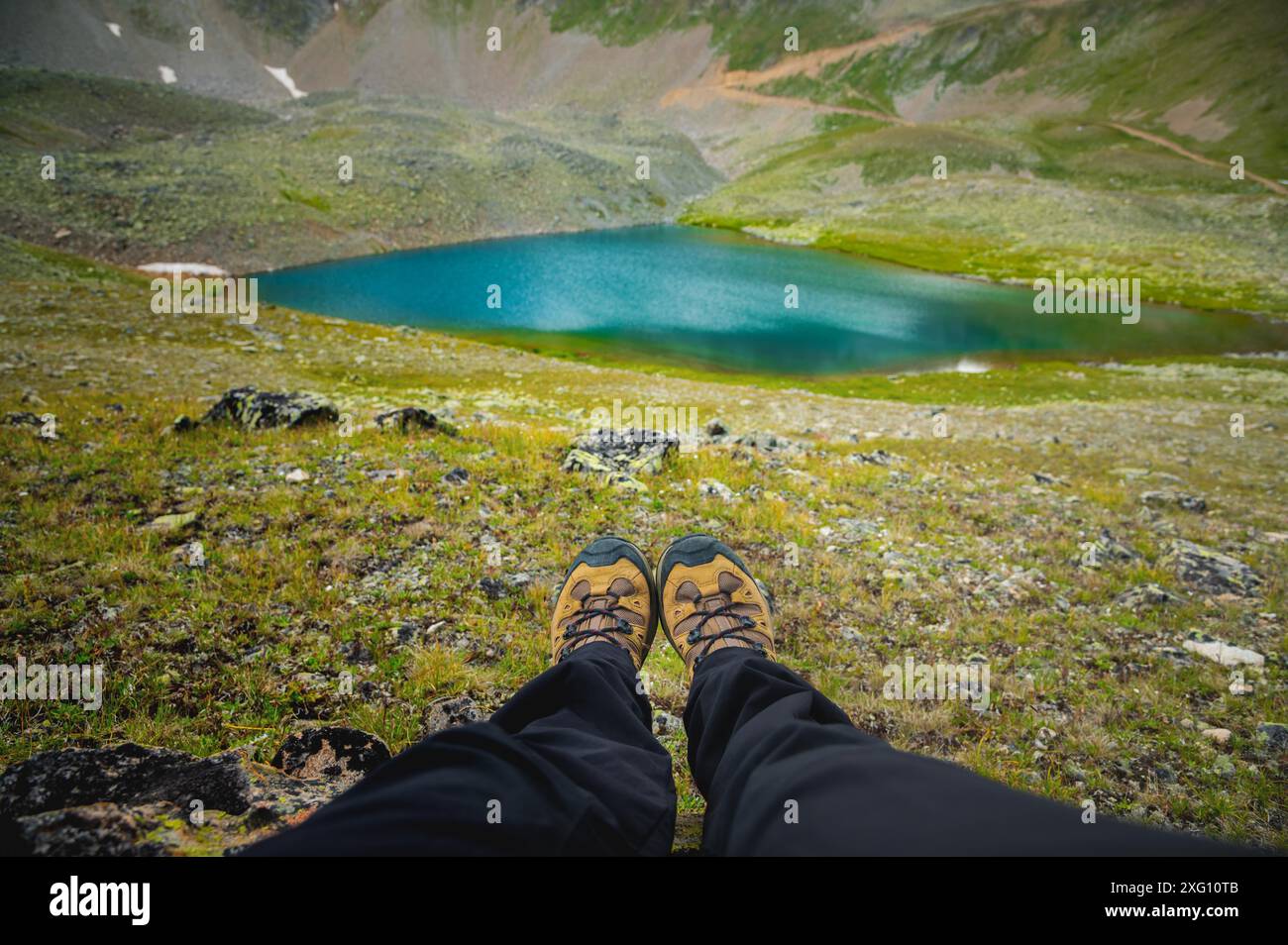 Il viaggiatore riposa su un altopiano di montagna. Vista in prima persona, gambe ravvicinate sullo sfondo di un lago turchese di montagna Foto Stock