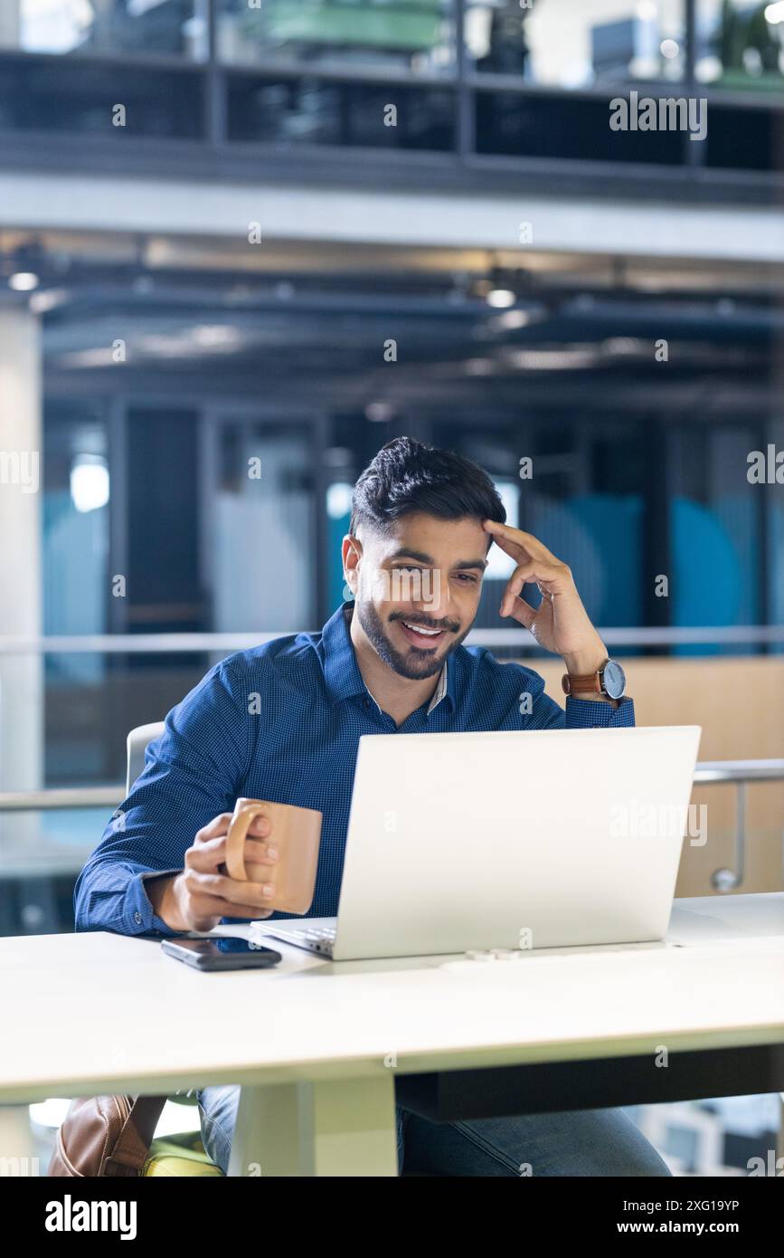 Un uomo d'affari sorridente che tiene in mano una tazza di caffè e utilizza un computer portatile in un ufficio moderno, spazio fotocopie Foto Stock