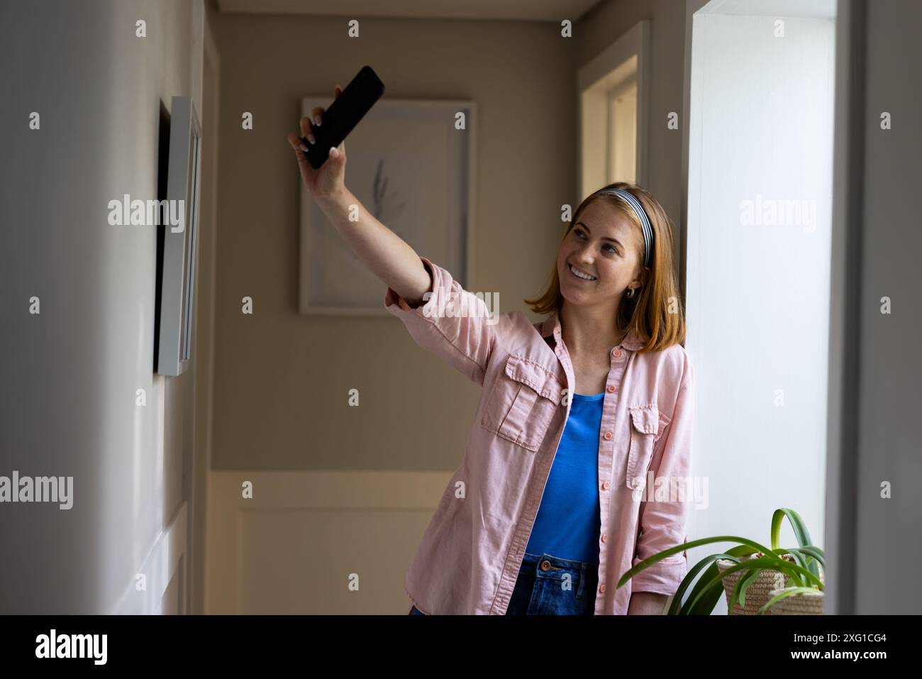 Scattare selfie, donna sorridente e tenere lo smartphone nel corridoio di casa Foto Stock