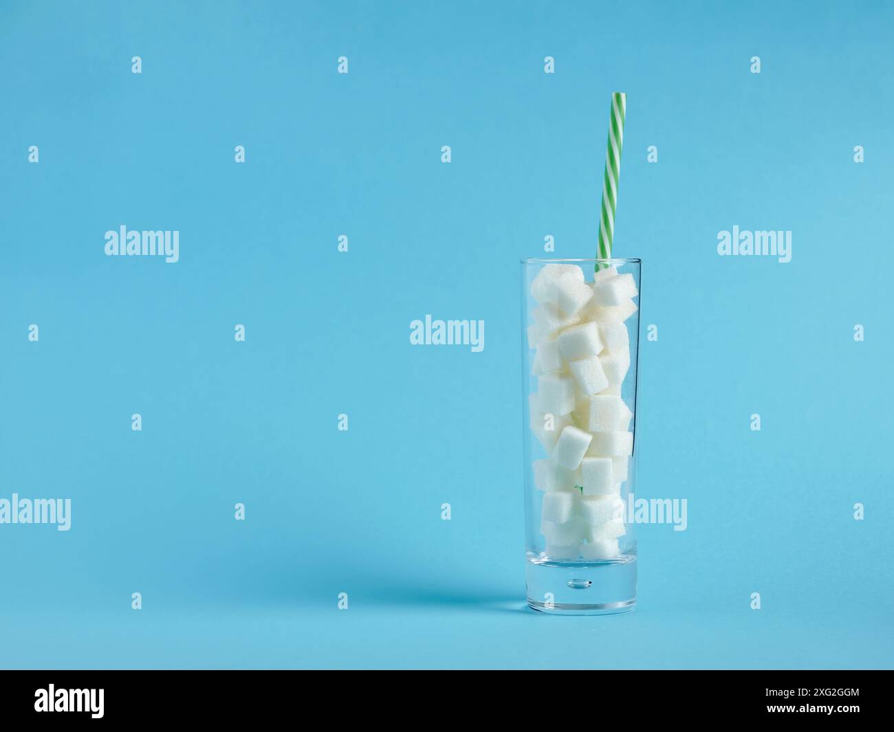 Consumo eccessivo di zucchero e dipendenza da glucosio. Pericoli di assunzione eccessiva di zucchero con le bevande. Bicchiere riempito con cubetti di zucchero e una cannuccia su un dorso blu Foto Stock