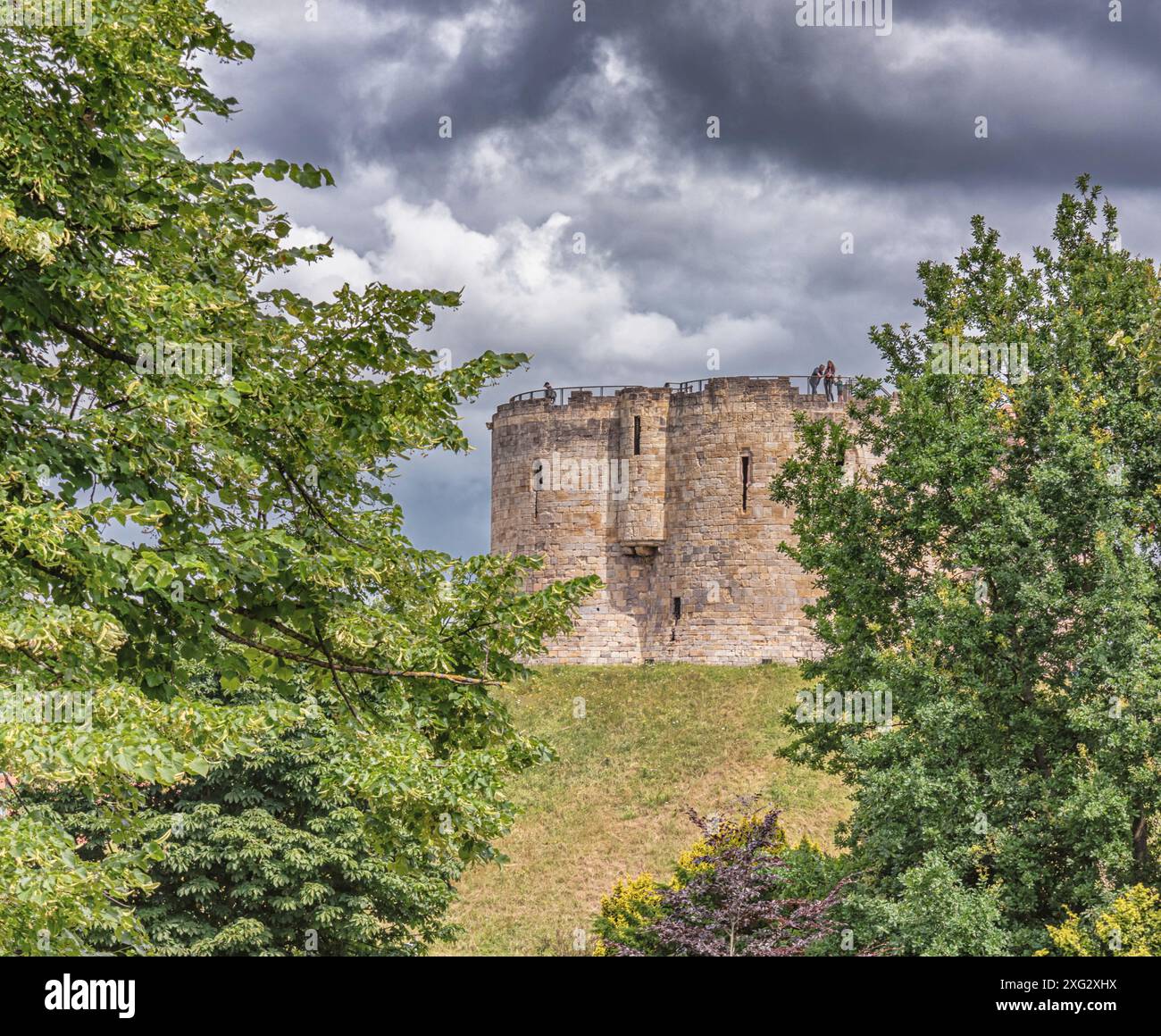 Un'antica torre del XIII secolo su un alto argine vista da due alberi. I turisti sono sulle pareti e sopra c'è un cielo nuvoloso. Foto Stock