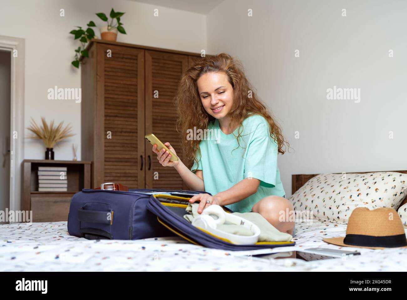 Una bruna riccia si siede sul letto e prepara le sue cose per le vacanze. Foto Stock