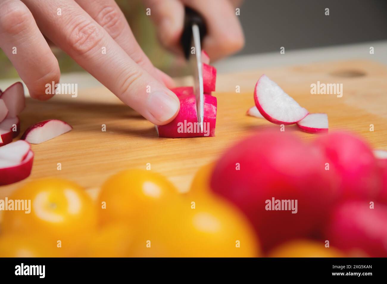 Primo piano di mani femminili tagliano il ravanello crudo fresco su una tavola di legno sullo sfondo di pomodori ciliegini gialli. Cibo vegetariano gustoso e salutare Foto Stock