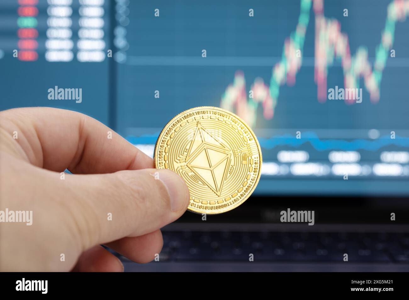 Mano che tiene una moneta di Ethereum e grafico sul display del laptop sullo sfondo. Concetto di criptovaluta o di investimento Foto Stock