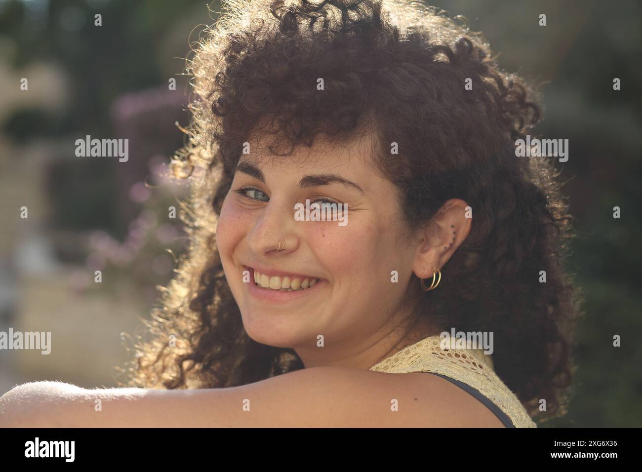 donna di 24 anni con capelli ricci, guarda la fotocamera con i suoi occhi azzurri mentre ride, il sole viene da dietro creando un bagliore da fotocamera. Foto Stock