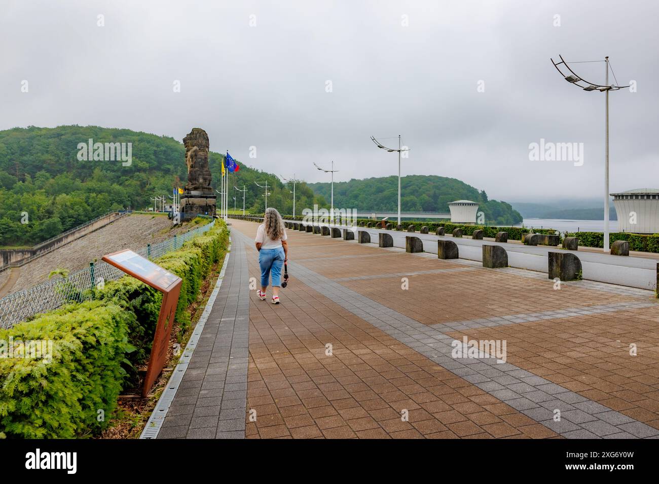 Jalhay, Belgio. 1° giugno 2024. Donna adulta anziana che cammina con la fotocamera sul sentiero pedonale in cima alla parete della diga di Gileppe, scultura di leoni e Foto Stock