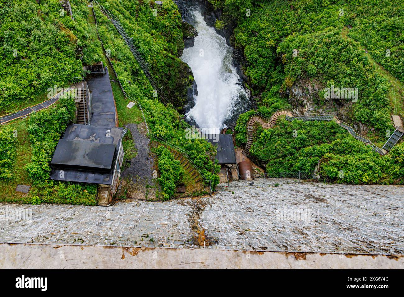 Prospettiva dall'angolo superiore dell'acqua che esce dallo spillway della diga di Robertville, a valle verso il fiume Warche che scorre tra i pendii rocciosi e la vegetazione selvaggia Foto Stock