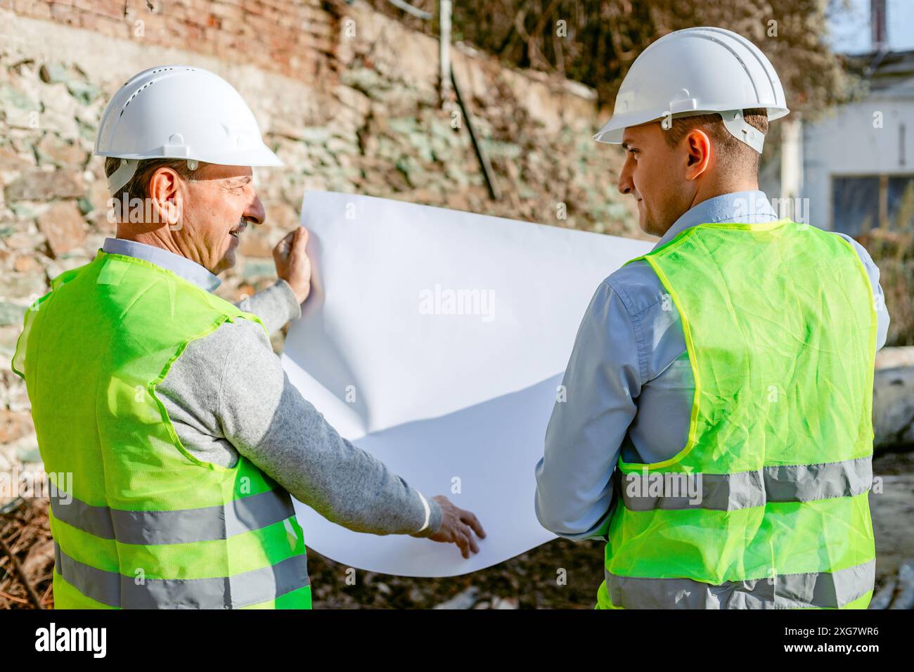 Due operai edili che indossano cappelli rigidi e giubbotti di sicurezza si trovano di fronte a una parete di mattoni in un cantiere edile. Foto Stock