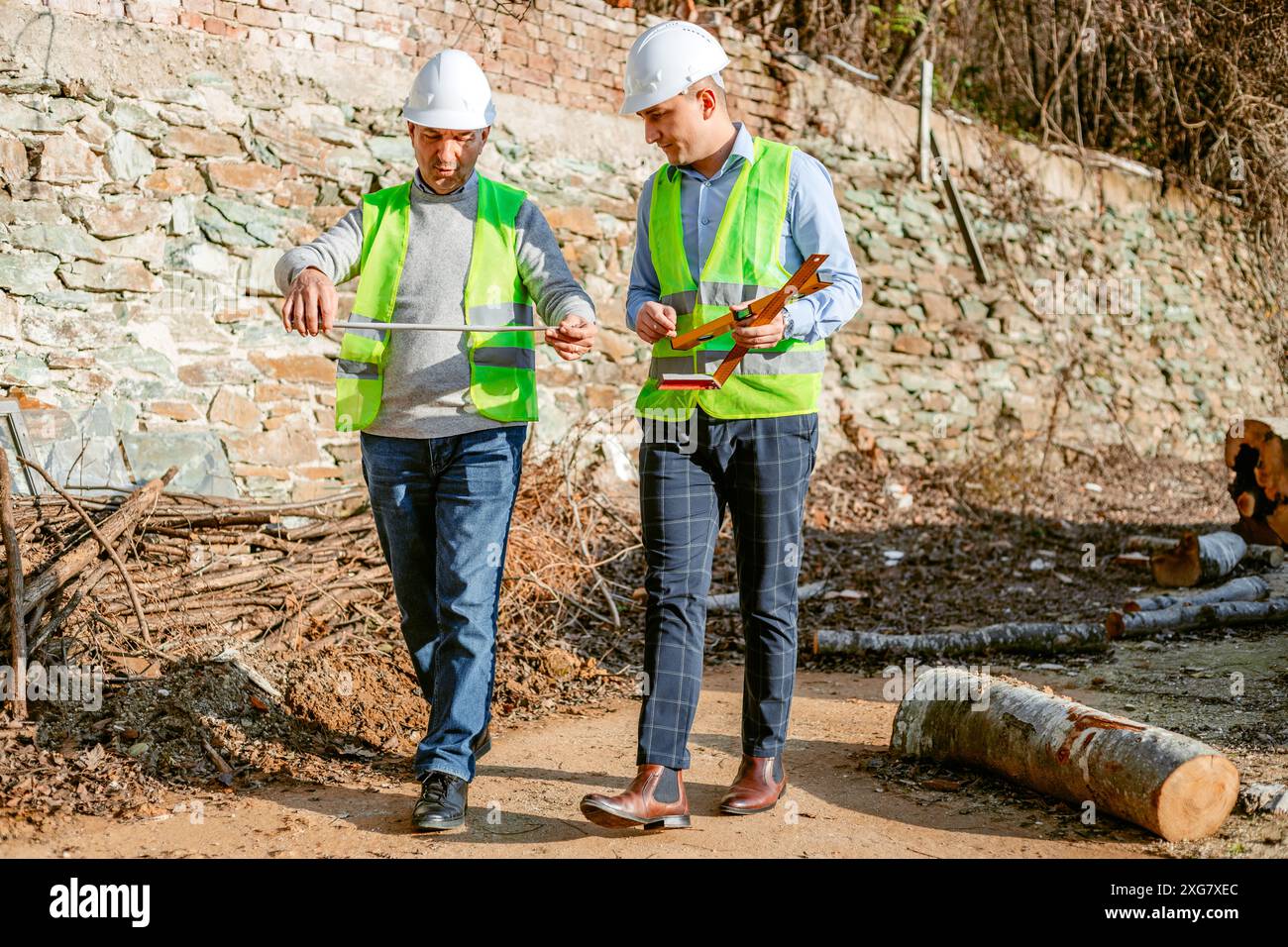 Due uomini che indossano cappelli rigidi e giubbotti di sicurezza attraversano una radura forestale. L'uomo a destra tiene un metro a nastro, mentre l'uomo a sinistra Foto Stock