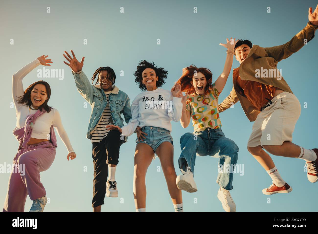 Un gruppo felice di amici che posa e salta insieme in una brillante giornata estiva sotto un cielo azzurro, abbracciando un divertimento spensierato. Foto Stock