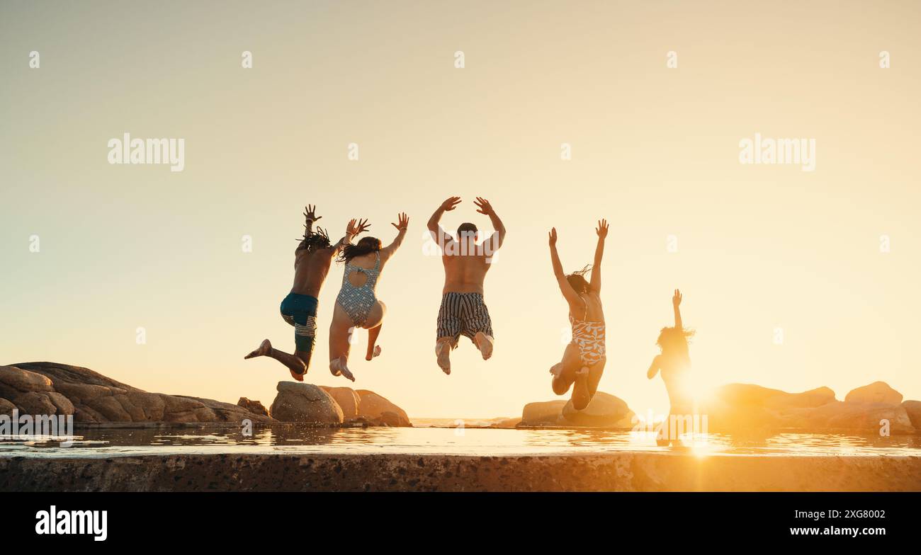 Gruppo di amici che saltano gioiosamente in acqua al tramonto, godendosi le loro vacanze estive all'estero. Viaggi e momenti di svago perfetti. Foto Stock
