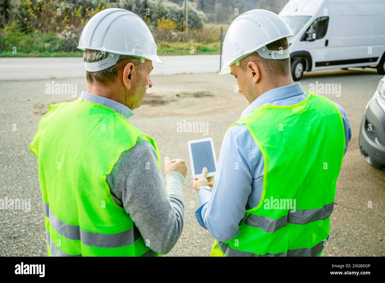 Due operai edili che indossano cappelli rigidi e giubbotti di sicurezza si trovano in un cantiere edile e rivedono i piani su un tablet. Foto Stock