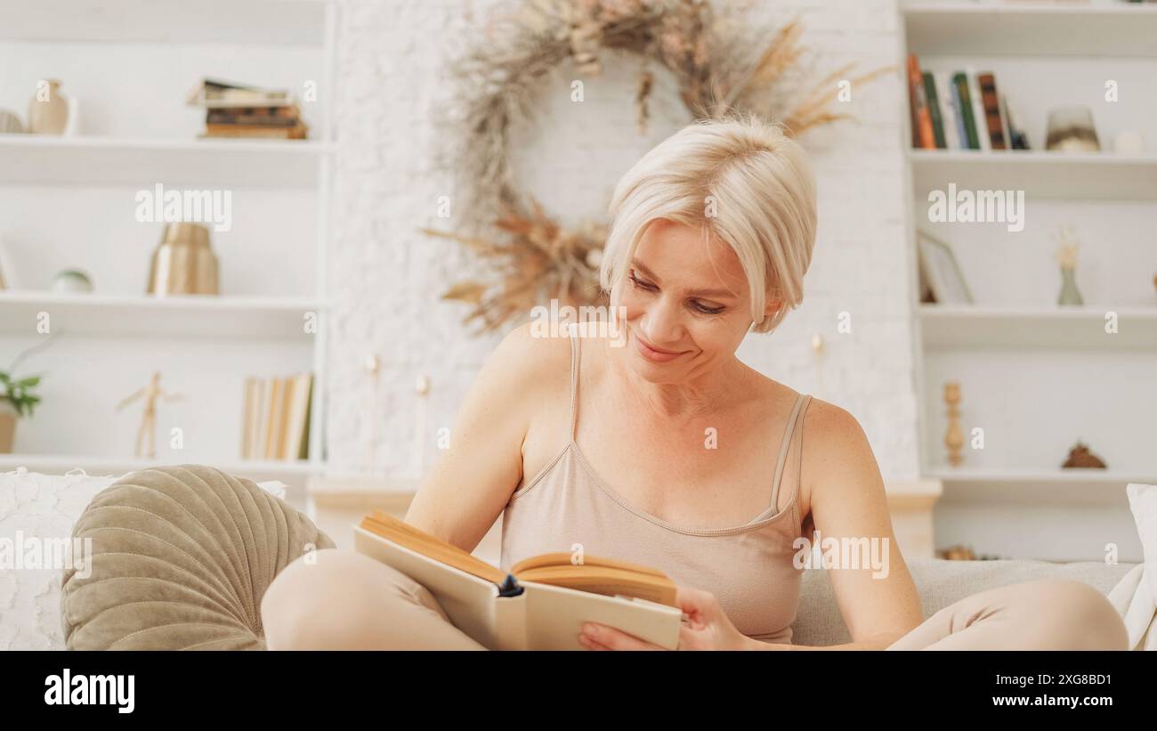 Libro interessante. Leggere gioia. Riposo domestico. Donna curiosa e felice in abiti per la casa eccitata ispirata da una storia inedita seduta su un divano in un luminoso salotto interno. Foto Stock