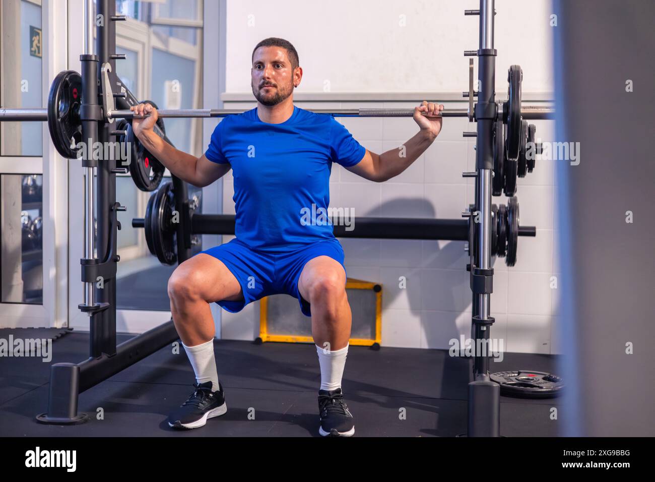 il giovane atleta di fitness è visto concentrato e determinato mentre esegue squat con pesi utilizzando una macchina multi-potenza durante la sua routine di allenamento in palestra. Foto Stock