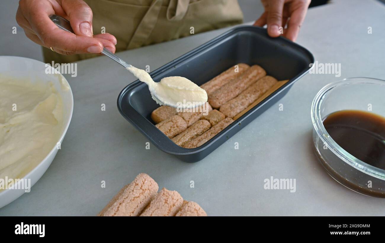 Primo piano di una persona che prepara il tiramisù con le savoiardi, la panna e il caffè. Preparazione passo passo del dessert catturata durante il processo. Foto Stock