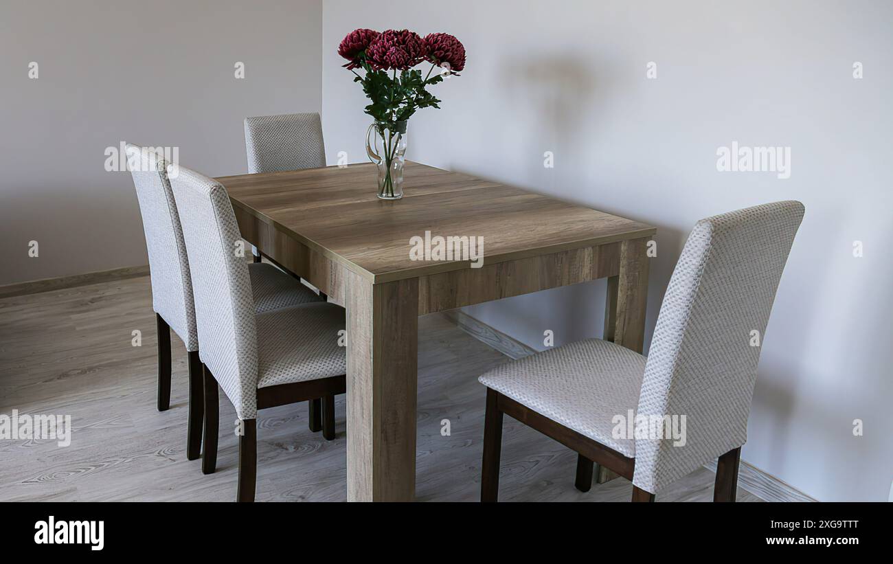 Un tavolo di legno marrone, quattro sedie rivestite in tessuto, un vaso di vetro con tre fiori rossi, pareti semplici grigie chiare e pavimento in laminato. Foto Stock