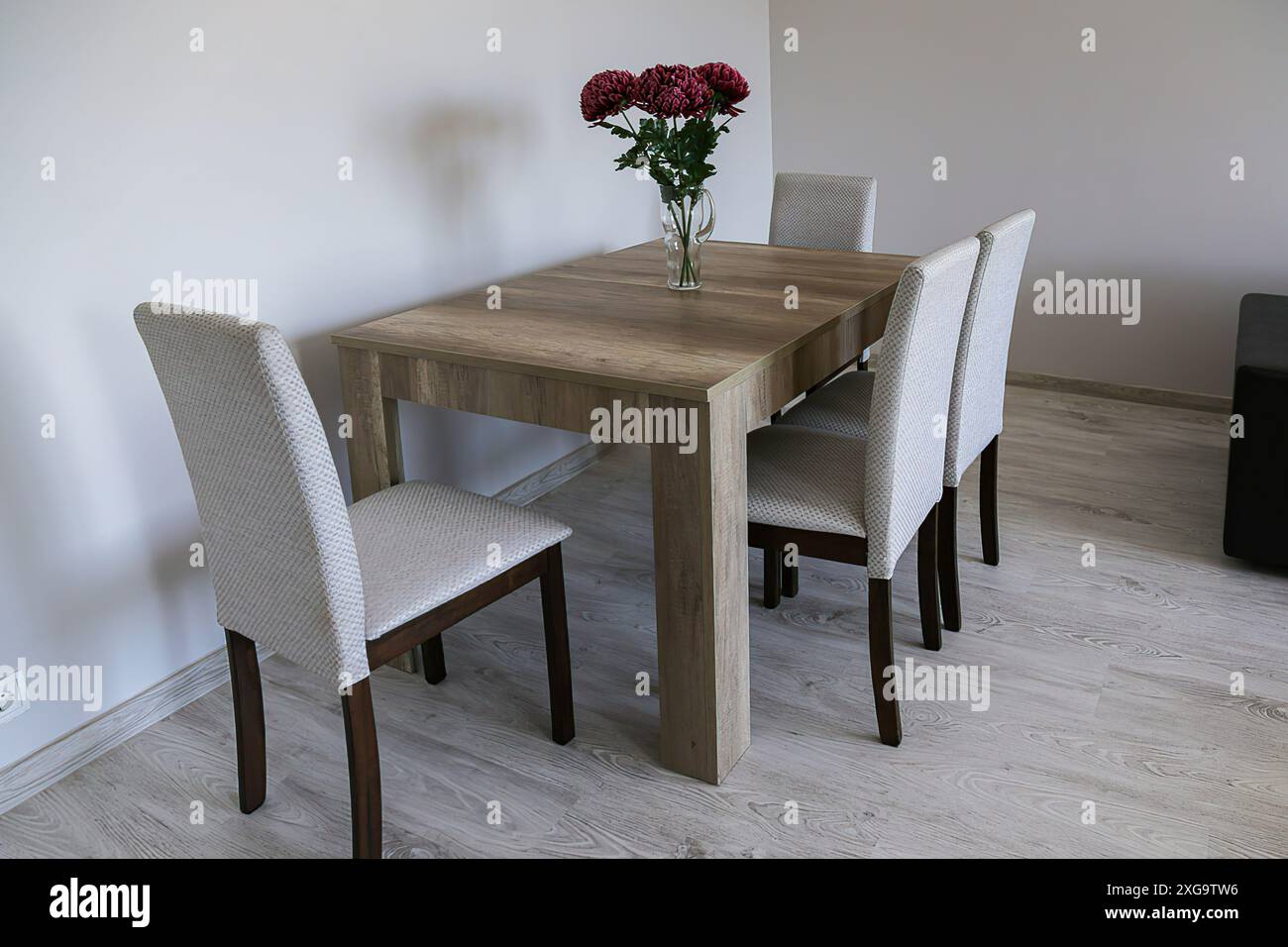 Un tavolo di legno marrone, quattro sedie chiare, un vaso di vetro con tre fiori rossi, pareti semplici grigie chiare e pavimento in laminato. Camera luminosa e accogliente. Foto Stock