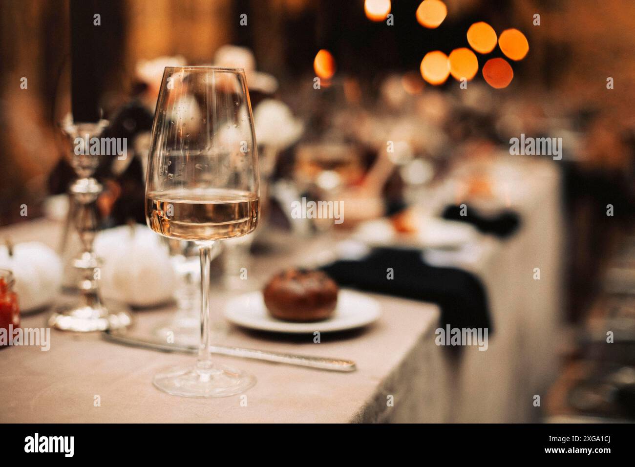 Focalizzazione selettiva sul vetro alto con vino bianco in primo piano, in piedi sullo sfondo sfocato del tavolo da pranzo non pulito dopo il banchetto festivo Foto Stock