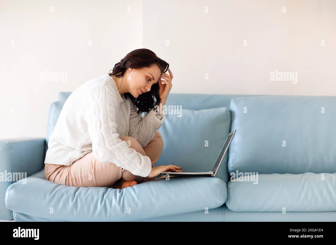 Giovane donna con un abbigliamento casual seduto con le gambe sul divano usando la tastiera del computer portatile, fissando lo schermo del computer, cercando importante Foto Stock