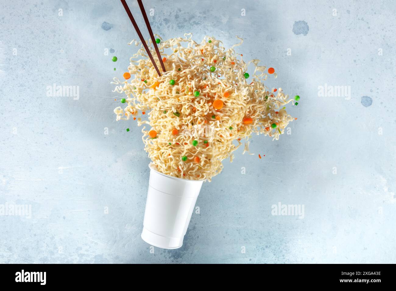 Tazza ramen con spaghetti istantanei che volano fuori da esso e bacchette, levitazione veloce del cibo. Piatto di noodle con carote e piselli verdi Foto Stock