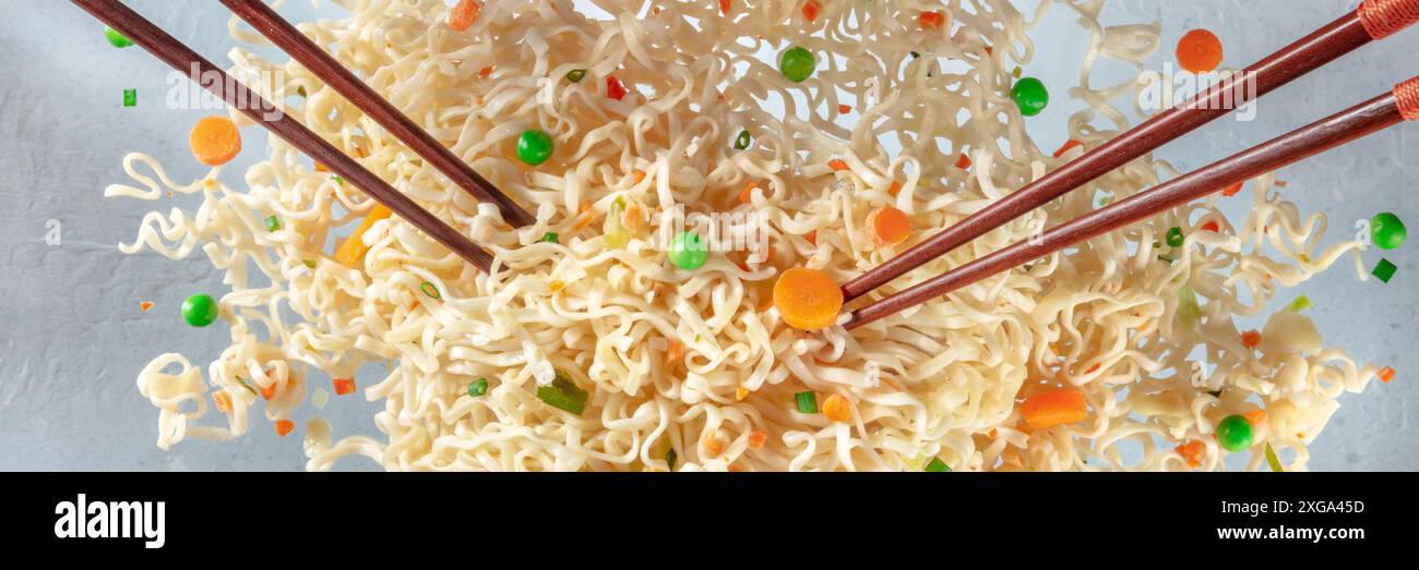Spaghetti volanti istantanei con due paia di panorama chopticks. Tazza ramen con carota e bandiera panoramica di piselli verdi Foto Stock