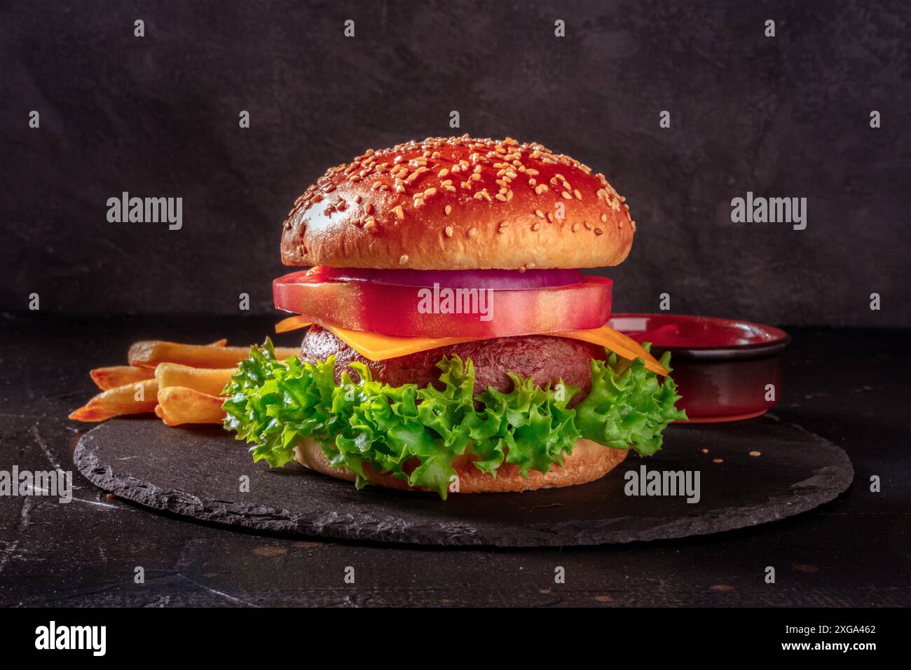 Vista laterale dell'hamburger sul nero, con patatine fritte e ketchup. Bistecca di manzo grassa con insalata verde, formaggio Cheddar, pomodoro di bistecca, cipolla viola e un Foto Stock