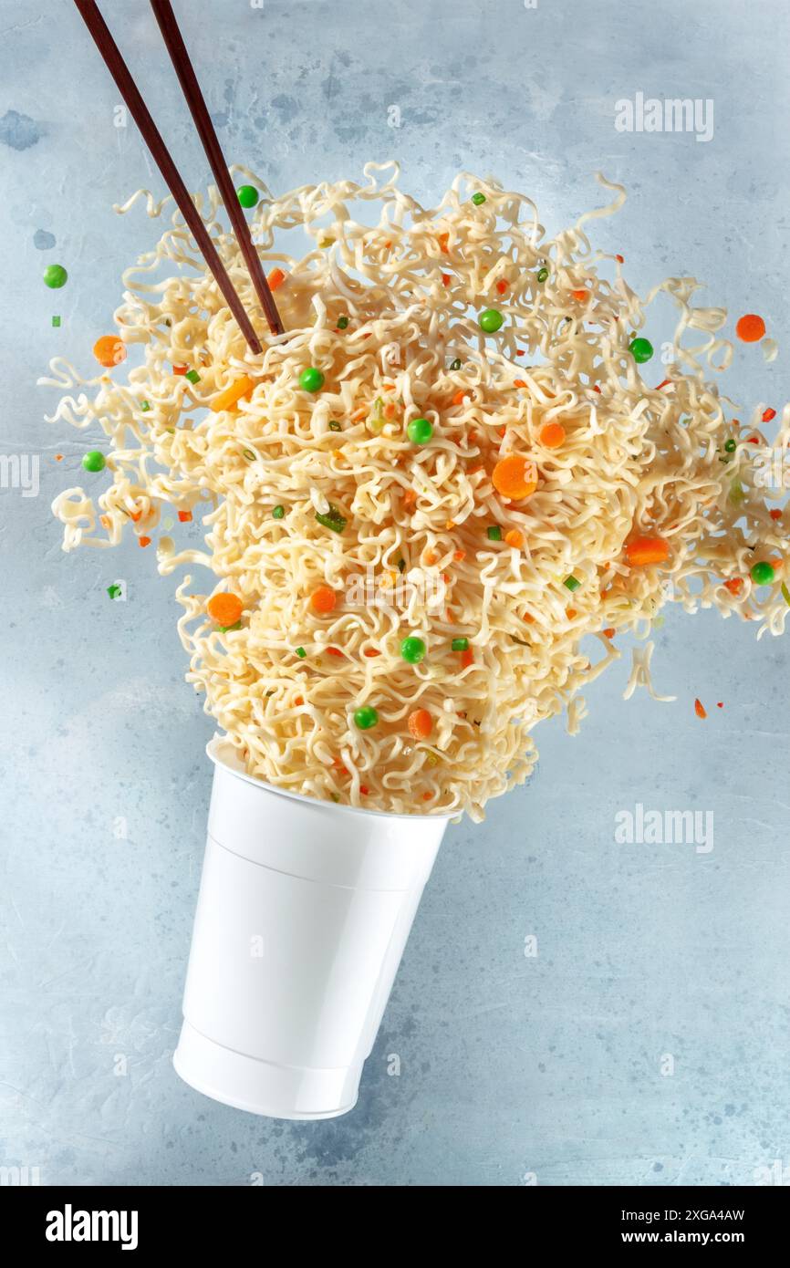 Tazza ramen con tagliatelle che galleggiano fuori di esso e bastoncini di chopsticks, levitazione veloce del cibo. Pasto istantaneo con carote e piselli verdi Foto Stock