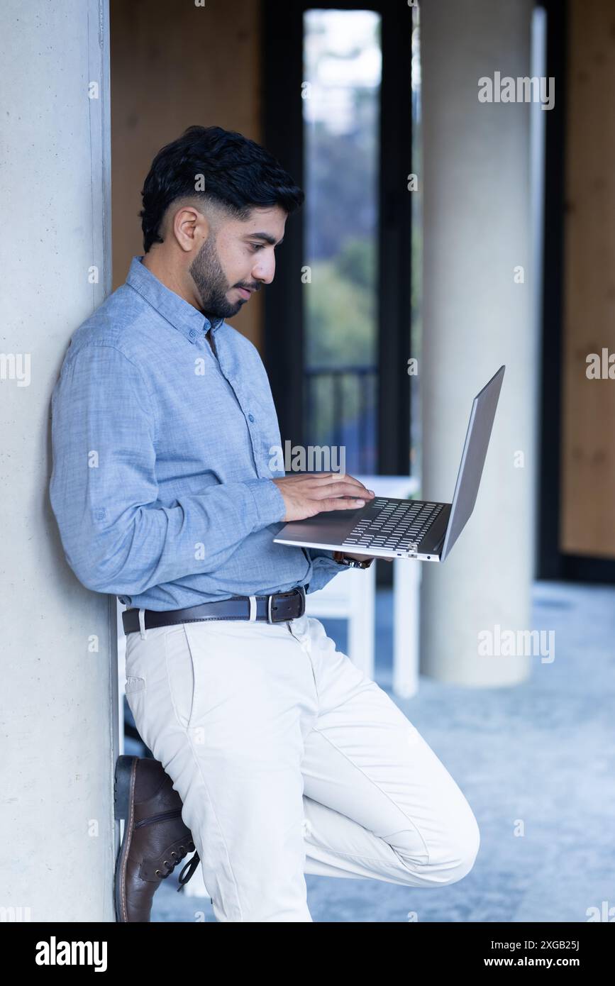Quando si utilizza un computer portatile, l'uomo si appoggia al muro in un moderno ufficio Foto Stock