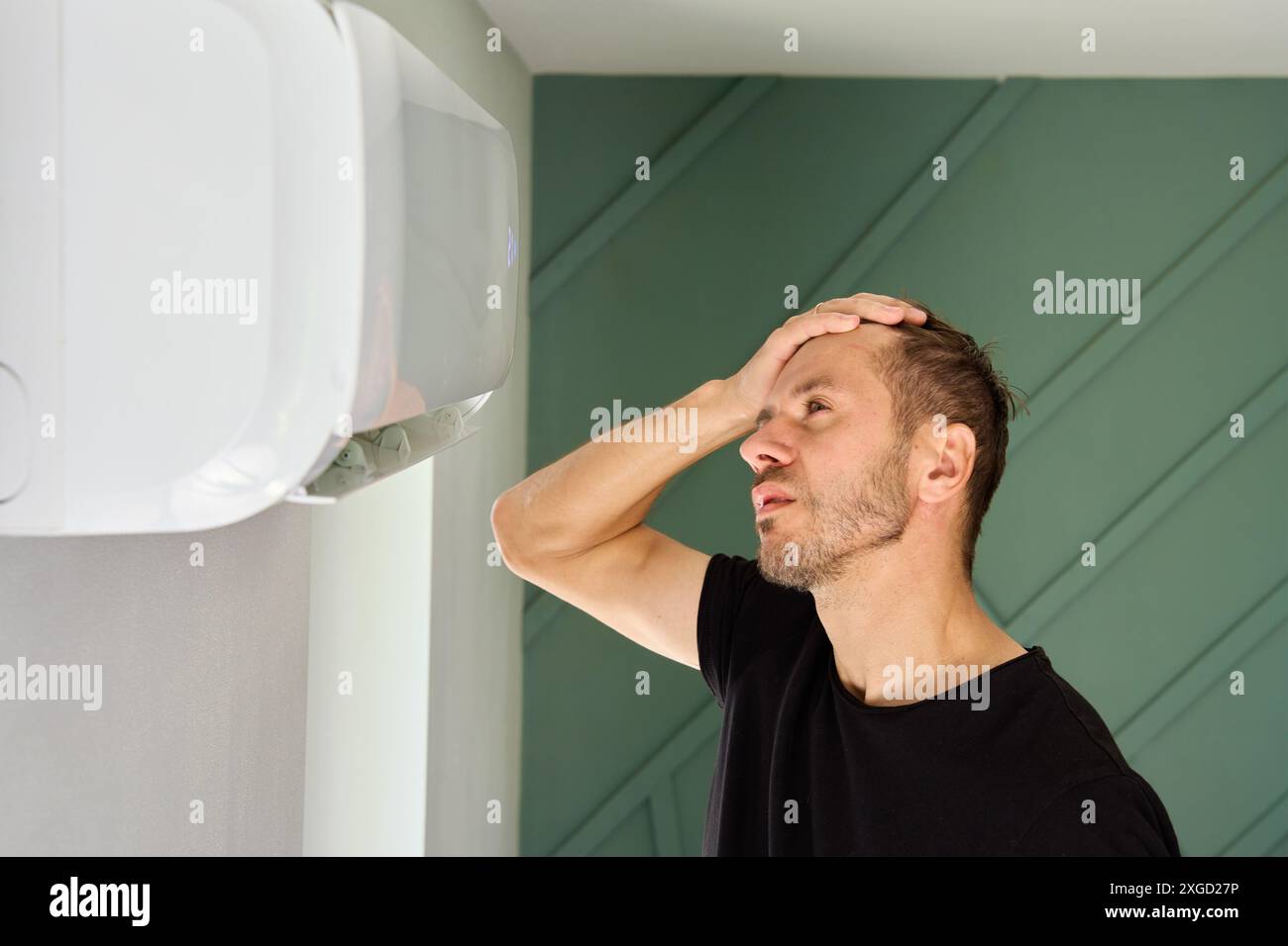 L'uomo si trova nel soggiorno accanto al condizionatore d'aria e gode di una brezza fresca. La persona sfugge dal calore scaricato sotto il condizionatore d'aria Foto Stock