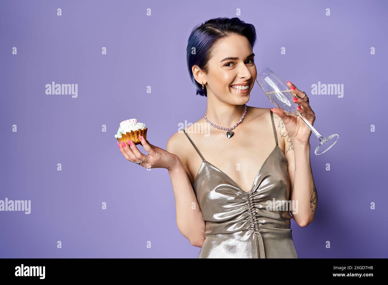 Una giovane donna elegante con i capelli azzurri corti in un abito argentato contiene un cupcake e un bicchiere di vino in una foto in studio. Foto Stock