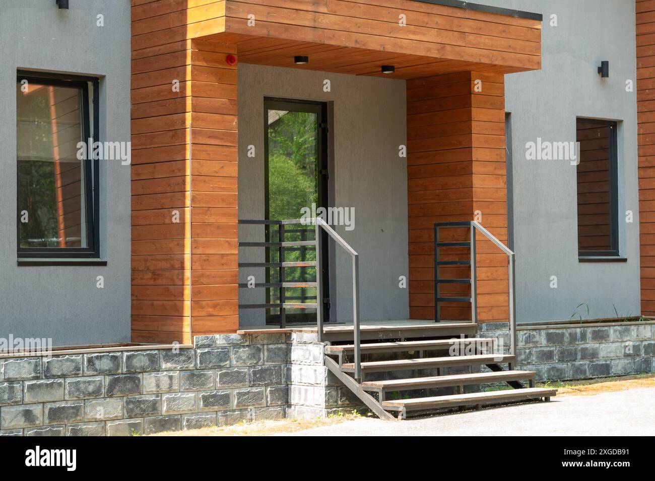 Una casa moderna esterna caratterizzata da un design elegante con rivestimenti in legno, accenti in pietra, una porta in vetro con telaio nero, scalini in cemento con ringhiera in metallo Foto Stock