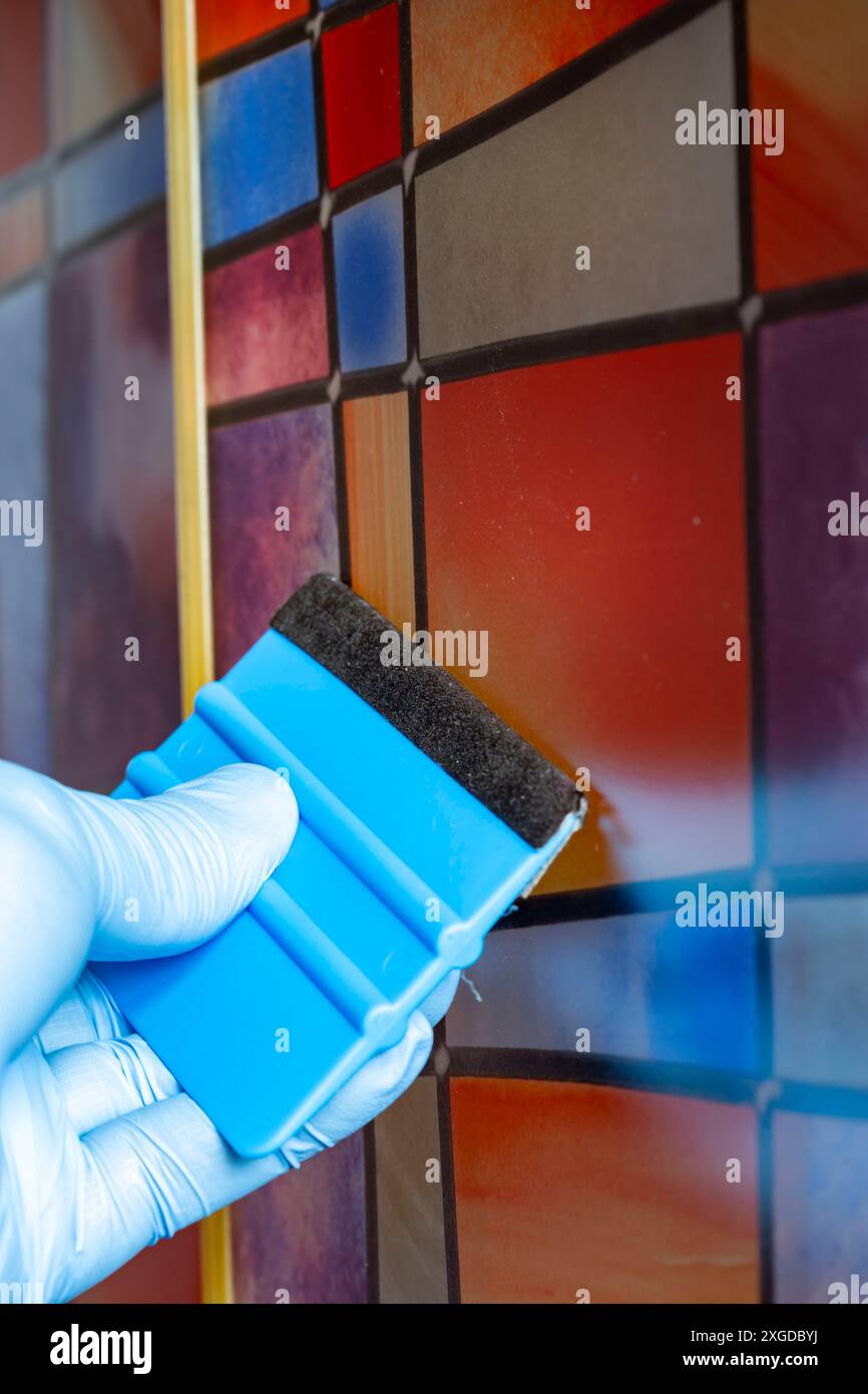 Persona che applica una pellicola di vetro colorato in plastica adesiva sul retro a uno schermo doccia in vetro. Levigare le bolle d'aria con un attrezzo. Foto Stock
