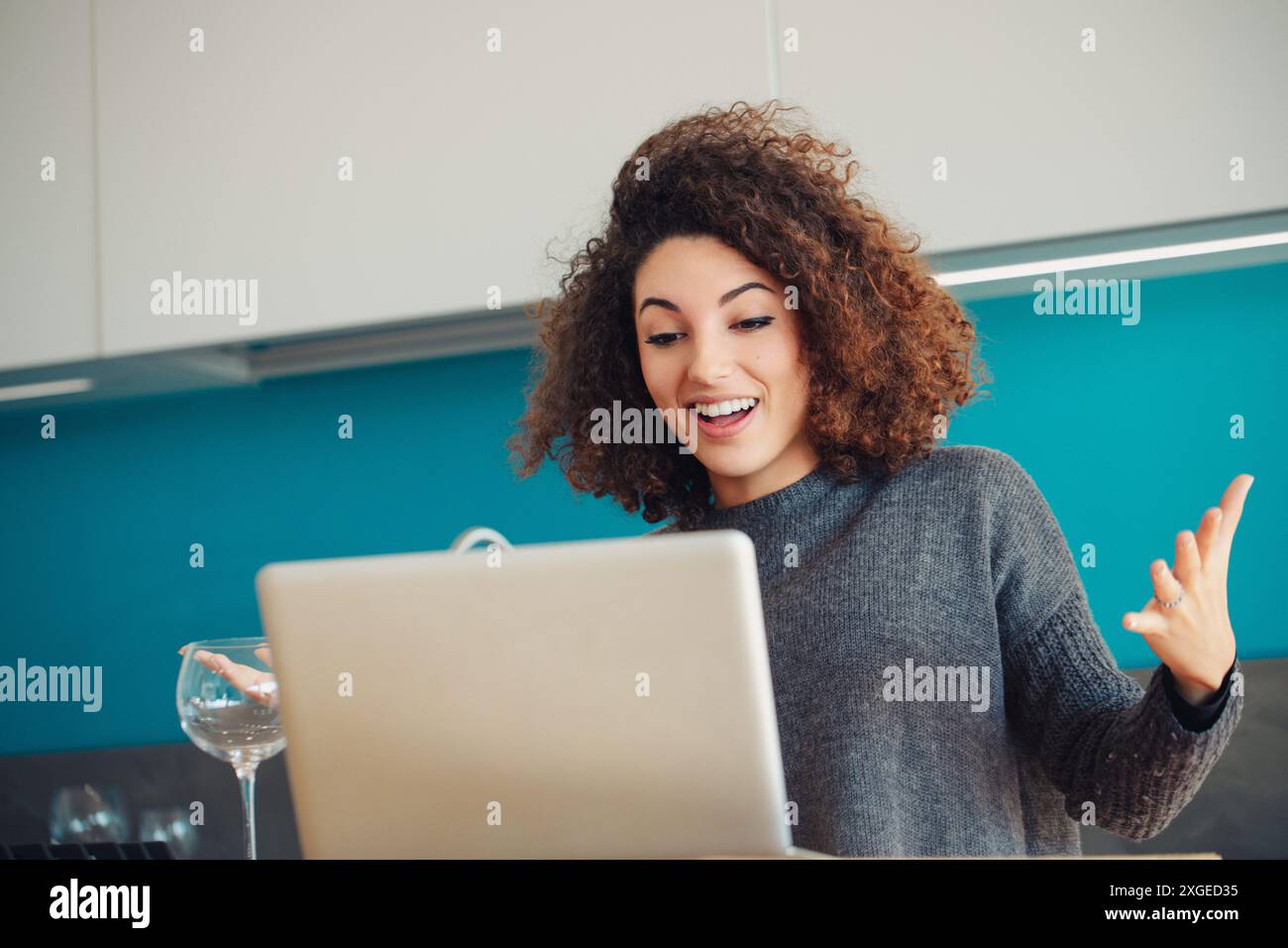 Una donna dai capelli ricci in cucina gestula durante una videochiamata, esprimendo la sua espressione vivace ed entusiasta. Il suo fidanzamento e il suo comportamento allegro suggeriscono Foto Stock