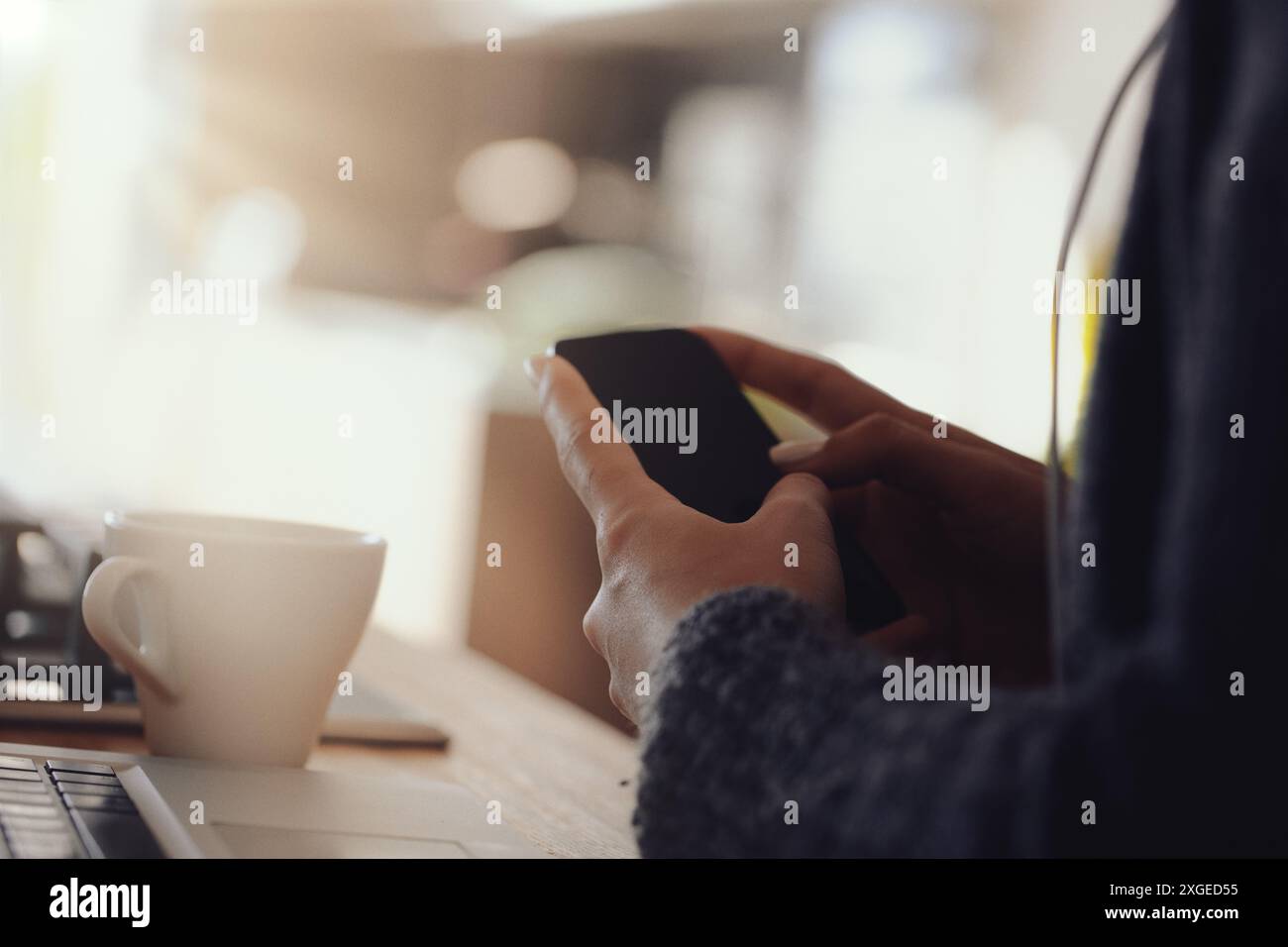 Le mani tengono uno smartphone con uno schermo vuoto in una stanza ben illuminata. Nelle vicinanze, una tazza da caffè bianca e altri oggetti su un tavolo di legno suggeriscono un ambiente tranquillo e informale Foto Stock
