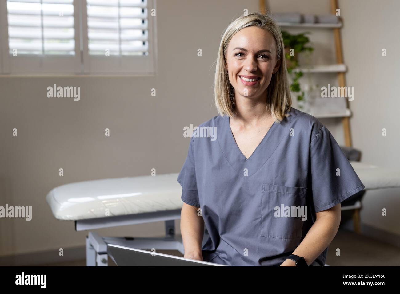 Donna sorridente in scrub seduto con laptop in clinica medica protesica, guardando fiducioso, copia sp Foto Stock