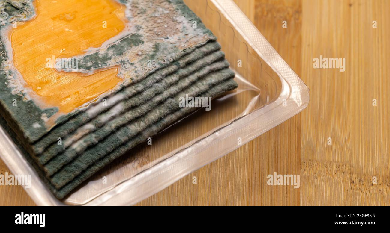 Fette di formaggio giallo ammuffito disposte su un tagliere di bambù. Spore di muffa verdi sul cibo. Cibo sprecato scaduto Foto Stock