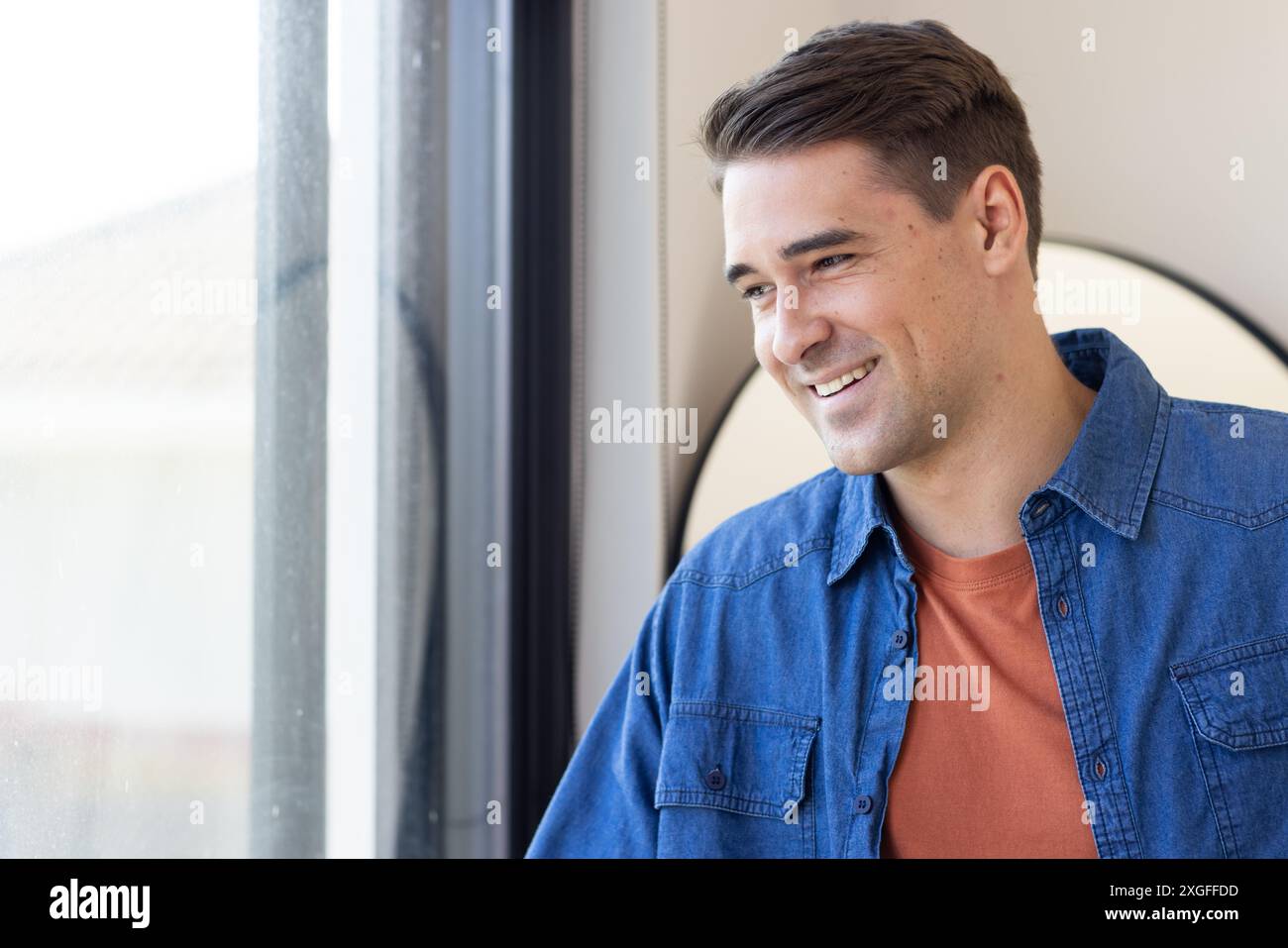 Uomo sorridente con camicia casual in denim in piedi accanto alla finestra, guardando all'esterno Foto Stock