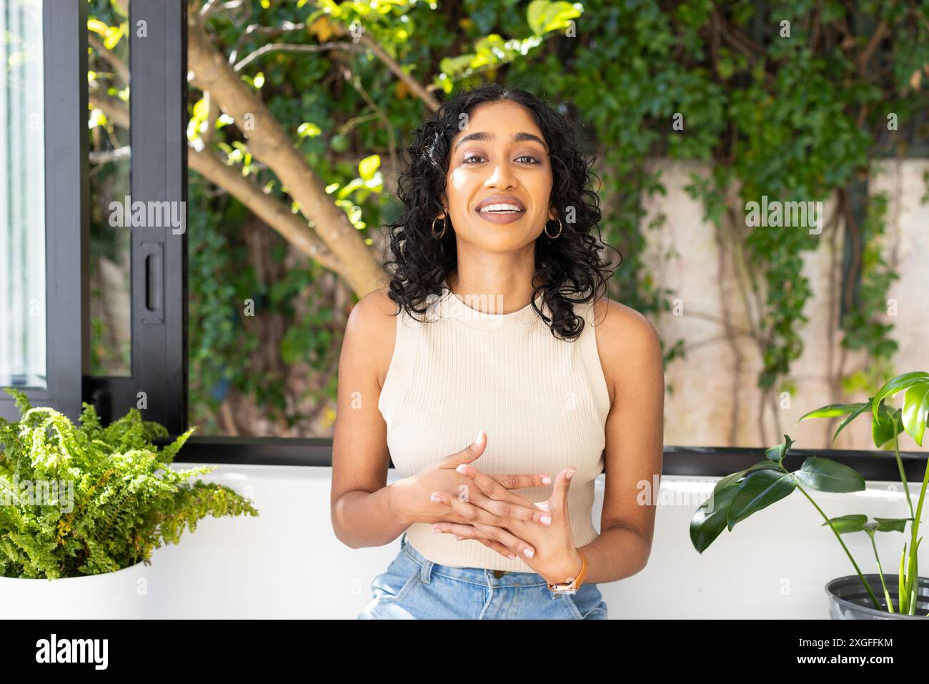 Donna sorridente in videochiamata in interni con piante, indossando abbigliamento casual e gioielli Foto Stock