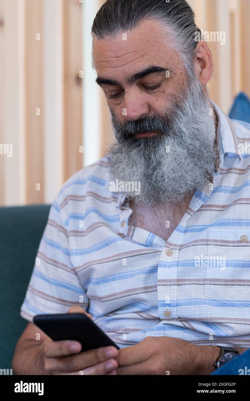 Utilizzando lo smartphone, un anziano barbuto che si concentra mentre si siede sul divano in casa Foto Stock