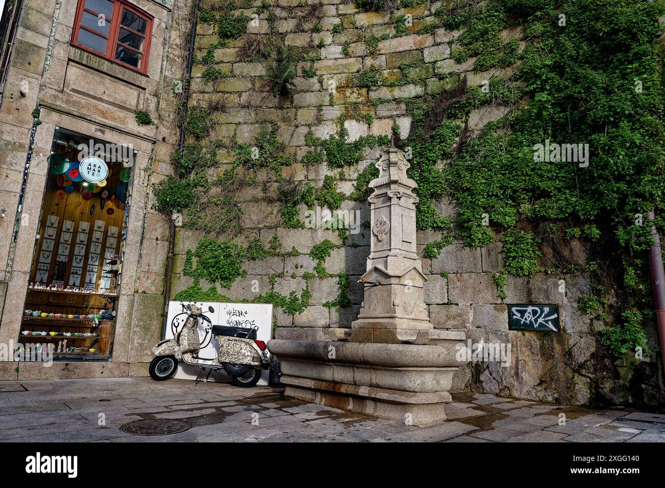 Angolo pittoresco di Porto con una vecchia fontana in pietra, un'affascinante esposizione di negozi e uno scooter d'epoca parcheggiato contro un muro coperto di viti Foto Stock