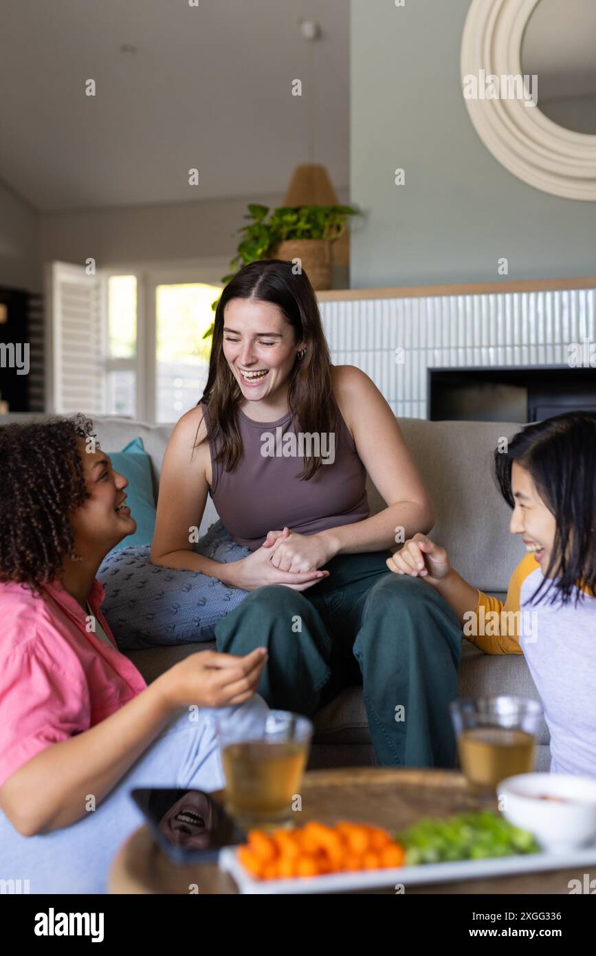 Ridendo e chiacchierando, tre donne amiche si godono spuntini e bevande a casa Foto Stock