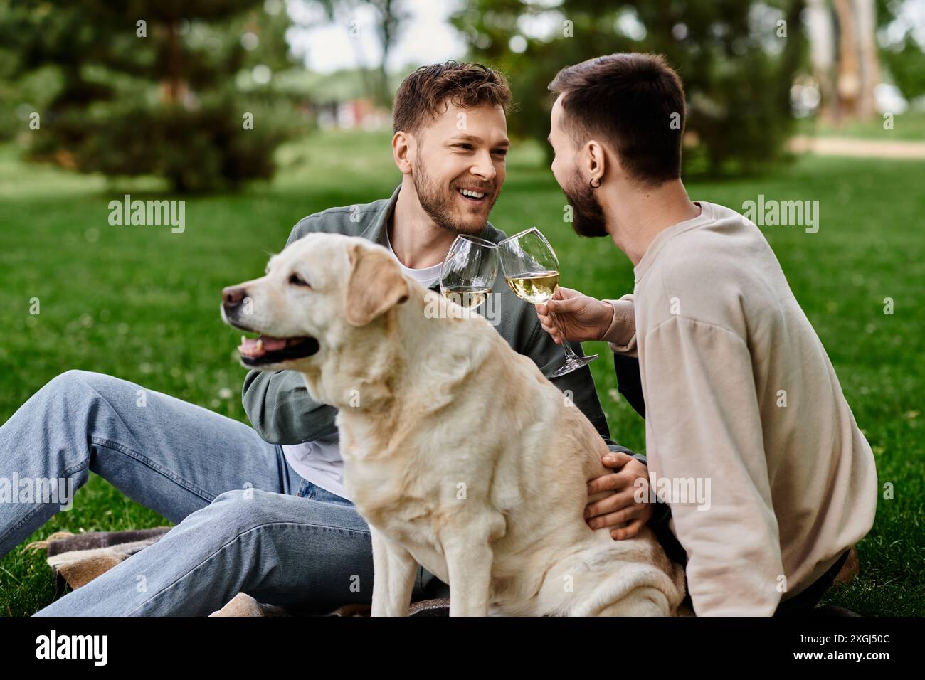 Una coppia gay con la barba si diverte a fare un picnic con il proprio cane labrador in un lussureggiante parco verde. Stanno sorridendo e ridendo, alzando i loro bicchieri di vino in una t Foto Stock